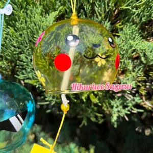 May include: A yellow glass wind chime with a cartoon face and red and yellow flower accents. The wind chime has a white string with yellow beads and the text "Hikariwo Sagasu" on the bottom.