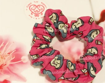 Op de afbeelding: Een roze stoffen scrunchie met een herhalend patroon van grijze cartoon gezichten en de tekst "Cassdoodles".