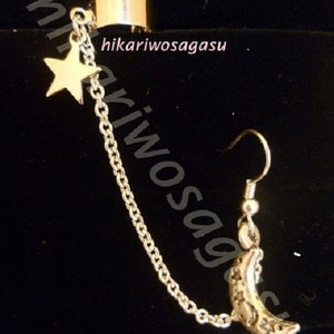 May include: Silver ear cuff with a dangling chain, a star charm, and a crescent moon charm. The chain is attached to the ear cuff and the charms dangle from the chain.