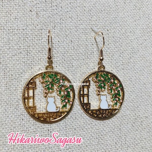 May include: Gold tone earrings with a white cat silhouette sitting in a window with green vines and pink flowers. The earrings are round and have a delicate design.