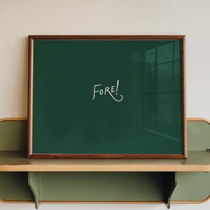 May include: A rectangular dark green print in a thin brown wooden frame featuring the white handwritten text "FORE!" in the center, resting on a wall-mounted olive green shelf with light wood trim and rounded edges.