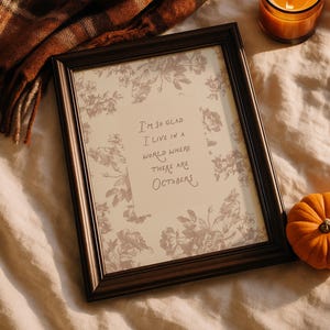 May include: A rectangular black picture frame containing a print with a grey floral pattern and the text I'm so glad I live in a world where there are Octobers. The frame is accompanied by a brown plaid blanket, a lit candle, and a small orange pumpkin.