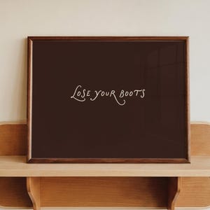 May include: A rectangular dark brown framed print featuring the text "LOSE YOUR BOOTS" in a light-colored, cursive-style font. The frame is made of dark wood and is positioned on a light-colored wooden shelf with rounded edges and side supports.
