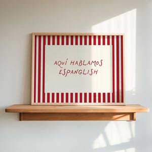 May include: A rectangular framed print featuring a cream-colored center with the red text "AQUÍ HABLAMOS ESPANGLISH" surrounded by a border of vertical red and white stripes. The artwork is enclosed in a light-toned wooden frame.