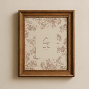 May include: A rectangular wooden picture frame with a dark brown finish containing a print. The print features a beige background with a repeating brown floral pattern and the centered text "How Lucky Are We" written in a simple, dark brown script.