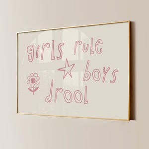May include: A rectangular wall art print in a thin gold frame featuring the pink hand-drawn text girls rule boys drool. The design includes a simple line-drawn star and a small flower icon on a light cream background, presented in a landscape orientation.