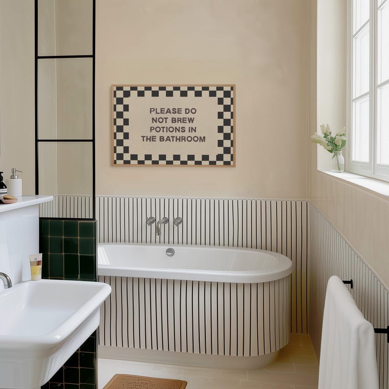 以下が含まれることがあります： 黒と白の市松模様の縁取りと、黒い大文字で「PLEASE DO NOT BREW POTIONS IN THE BATHROOM」というテキストが書かれた長方形のウォールアートプリント。明るい木製のフレームに入れられ、白いバスタブの上のベージュの壁に飾られている。