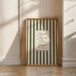 Może przedstawiać: Oprawiony wydruk artystyczny ze złotą ramą. Grafika przedstawia zielono-białe paski z napisem "Meet Me On The Green" w odręcznej czcionce. Wydruk jest wyświetlany na białej ścianie.