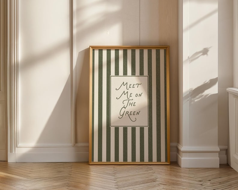 Może przedstawiać: Oprawiony wydruk artystyczny ze złotą ramą. Grafika przedstawia zielono-białe paski z napisem "Meet Me On The Green" w odręcznej czcionce. Wydruk jest wyświetlany na białej ścianie.