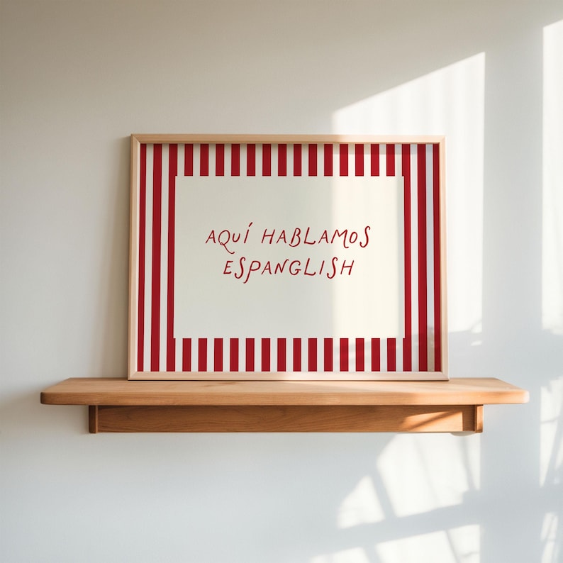 May include: A framed print with a red and white striped border and the text "AQUÍ HABLAMOS ESPANGLISH" in red. The print is displayed on a wooden shelf against a white wall.