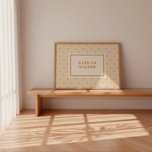 Puede incluir: Una pieza de arte mural rectangular enmarcada con un fondo crema claro y un patr&oacute;n repetitivo de peque&ntilde;os puntos dorados. Centrado dentro de un borde rectangular dorado se encuentra el texto "LIFE IS GOLDEN" en una fuente sans-serif dorada.