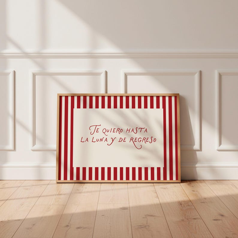 May include: A framed print with a cream-colored center and a red and white striped border. The print features the Spanish text "Te quiero hasta la luna y de regreso" in red script, set against a light wood frame.