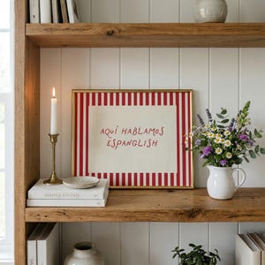 May include: A rectangular framed print with a red and white vertical striped border and a cream centre featuring the red text "AQU&Iacute; HABLAMOS ESPANGLISH". The print is displayed on a wooden shelf next to a white candle and a bouquet of flowers.