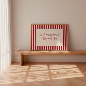 May include: A rectangular framed print featuring a white centre with the red text "AQU&Iacute; HABLAMOS ESPANGLISH" surrounded by a border of vertical red and white stripes. The artwork is displayed in a light-coloured wooden frame with a clean design.