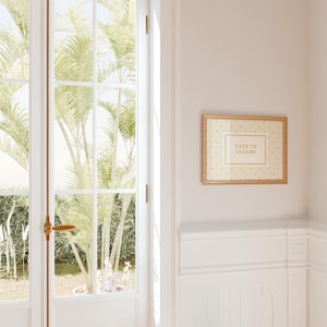Puede incluir: Una pieza de arte mural rectangular en un marco de madera clara con un fondo color crema, un patr&oacute;n de peque&ntilde;os puntos dorados y un borde rectangular central que contiene el texto "LIFE IS GOLDEN" en una fuente dorada sin remates.