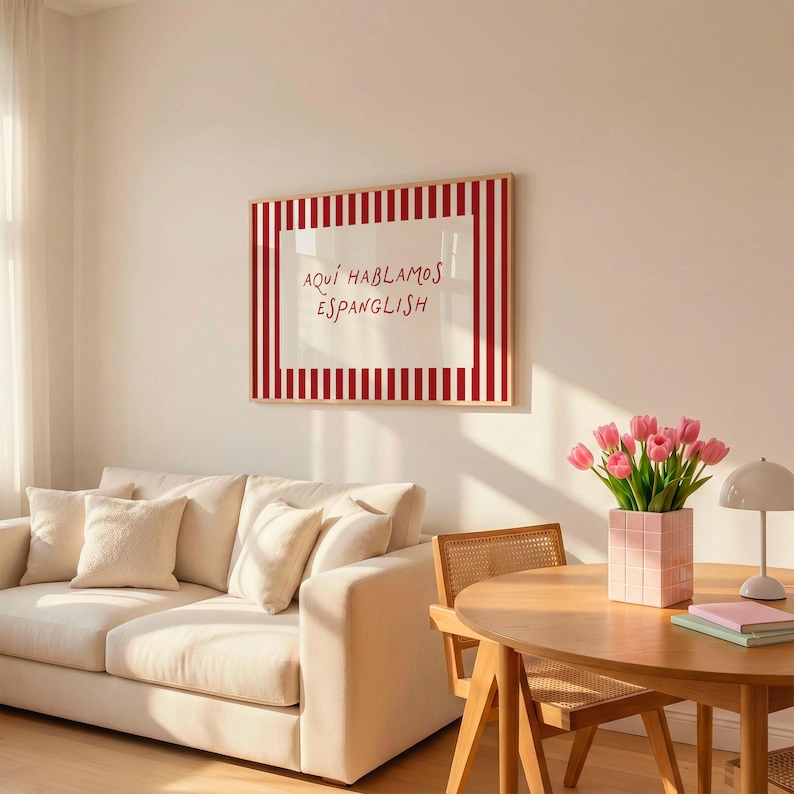 May include: A rectangular wall art print in a light wood frame featuring a white centre with the red text "AQU&Iacute; HABLAMOS ESPANGLISH" surrounded by a red and white vertical striped border, displayed on a plain wall above a cream-coloured sofa.