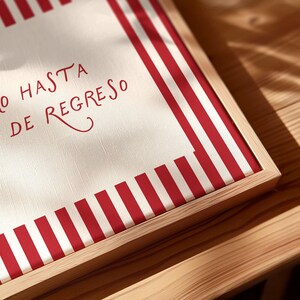 May include: A framed canvas with a red and white striped border. The canvas has the words "O HASTA DE REGRESO" in red script. The frame is made of light-colored wood, and the piece is set on a wooden surface.