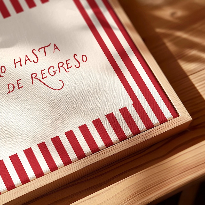 May include: A framed canvas with a red and white striped border. The canvas has the words "O HASTA DE REGRESO" in red script. The frame is made of light-colored wood, and the piece is set on a wooden surface.