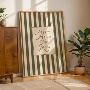 Może przedstawiać: Oprawiony wydruk ścienny z zielono-beżowym wzorem w paski. Grafika zawiera tekst "Meet Me On The Green" w ozdobnej czcionce. Wydruk opiera się o ścianę obok drewnianej szafki i roślin doniczkowych.