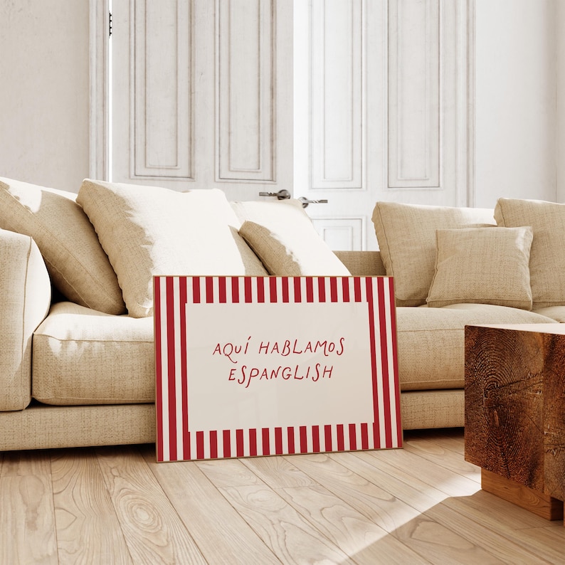 May include: A rectangular sign featuring a white centre panel with the red text "AQU&Iacute; HABLAMOS ESPANGLISH" surrounded by a border of vertical red and white stripes. The sign is positioned upright on a light-coloured wooden floor in front of a sofa.