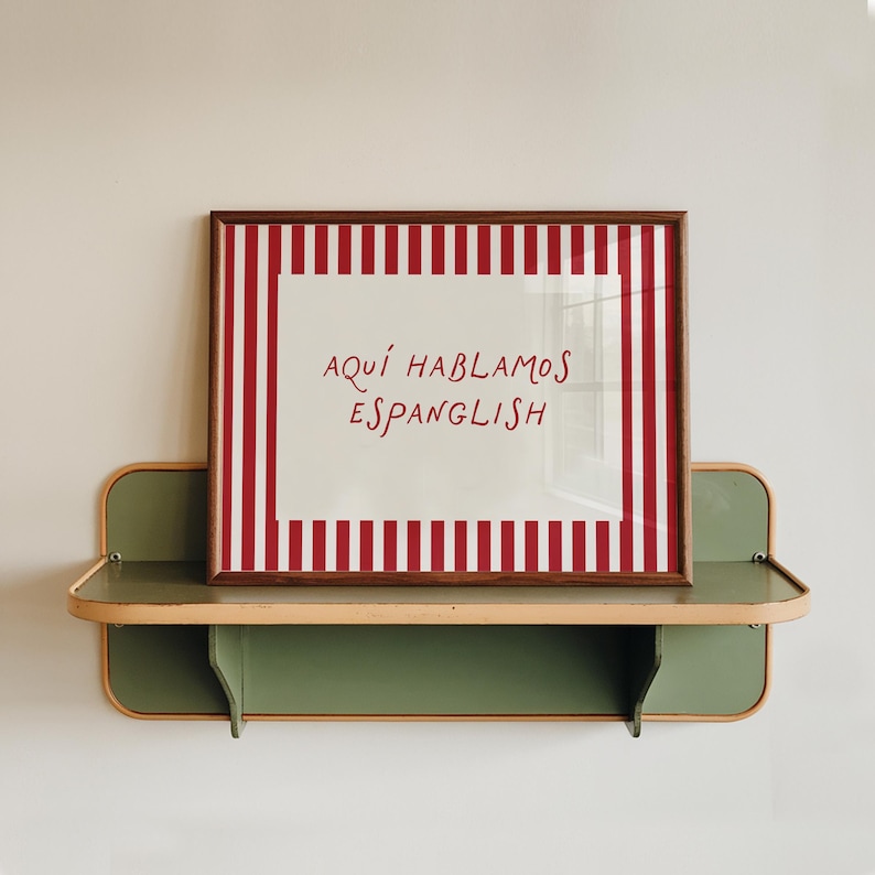 May include: A rectangular framed print featuring a white centre with the red text "AQU&Iacute; HABLAMOS ESPANGLISH" surrounded by a red and white vertical striped border. The print is displayed on a sage green wall-mounted shelf with light wood trim.