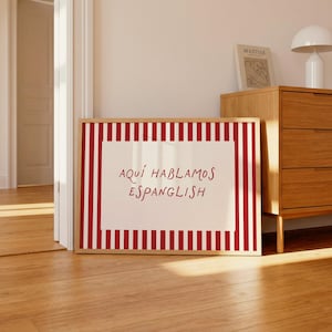 May include: A rectangular framed print featuring a white central panel with the red text "AQU&Iacute; HABLAMOS ESPANGLISH" surrounded by a border of vertical red and white stripes. The artwork is encased in a light-coloured wooden frame.