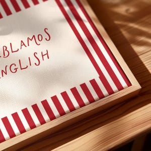 May include: A square canvas print in a light wood frame featuring a white background with vertical red stripes along the edges. The text "HABLAMOS ENGLISH" is printed in red, handwritten-style lettering on the left side of the canvas surface.