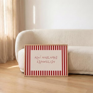 May include: A rectangular framed print featuring a cream-coloured centre with the red text "AQU&Iacute; HABLAMOS ESPANGLISH" surrounded by a border of vertical red and white stripes. The artwork is displayed in a light-toned wooden frame.