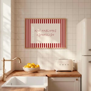 May include: A rectangular wall art print in a light wood frame featuring a cream-coloured centre with the red text "AQU&Iacute; HABLAMOS ESPANGLISH" surrounded by a border of vertical red and white stripes, displayed on a white tiled kitchen wall.