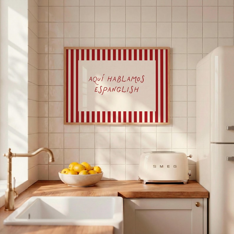 May include: A rectangular wall art print in a light wood frame featuring a cream-coloured centre with the red text "AQU&Iacute; HABLAMOS ESPANGLISH" surrounded by a border of vertical red and white stripes, displayed on a white tiled kitchen wall.