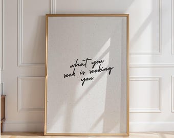 May include: A framed print with the text "what you seek is seeking you" in a handwritten style. The frame is gold, and the background is a light beige color. The print is leaning against a white wall.