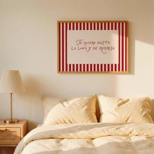 May include: A framed print with red and white vertical stripes and the text "Te quiero hasta la luna y de regreso" in red script. The print hangs above a bed with yellow bedding and pillows. A lamp with a pleated shade sits on a nightstand.