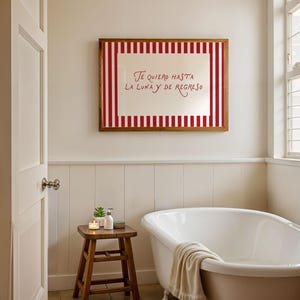 May include: A bathroom scene featuring a white bathtub, a wooden stool with toiletries, and a framed artwork. The artwork has a red and white striped border and the text "Te quiero hasta la luna y de regreso". The room has white walls and a window.