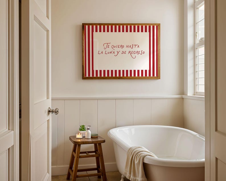 May include: A bathroom scene featuring a white bathtub, a wooden stool with toiletries, and a framed artwork. The artwork has a red and white striped border and the text "Te quiero hasta la luna y de regreso". The room has white walls and a window.