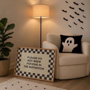 以下が含まれることがあります： 黒と白の市松模様の縁取りと「PLEASE DO NOT BREW POTIONS IN THE BATHROOM」というテキストが書かれた長方形の額入りサイン。このサインは、お化けの模様が入った黒いクッションが置かれたクリーム色の肘掛け椅子の横の床に置かれています。