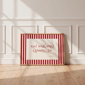 May include: A rectangular framed print featuring a cream-coloured centre with the red text 'AQU&Iacute; HABLAMOS ESPANGLISH' surrounded by a border of vertical red and white stripes. The frame is made of light-toned wood and has a clean, simple design.