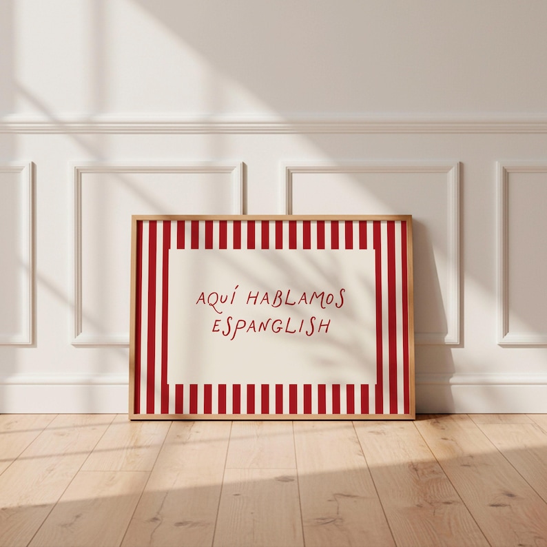 May include: A rectangular framed print featuring a cream-coloured centre with the red text 'AQU&Iacute; HABLAMOS ESPANGLISH' surrounded by a border of vertical red and white stripes. The frame is made of light-toned wood and has a clean, simple design.