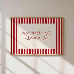 May include: A rectangular wall art print in a light wood frame featuring a white centre panel with the red text 'AQU&Iacute; HABLAMOS ESPANGLISH'. The border consists of vertical red and white stripes surrounding the central text area.