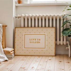 Puede incluir: Una impresi&oacute;n rectangular enmarcada con un fondo crema y un patr&oacute;n repetitivo de peque&ntilde;os puntos dorados. El centro contiene un rect&aacute;ngulo con borde dorado y el texto "LIFE IS GOLDEN" impreso en una tipograf&iacute;a dorada, negrita y sin remates.