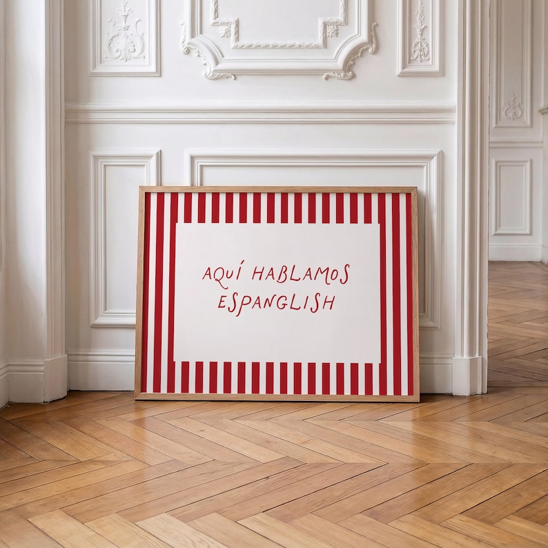 May include: A rectangular framed print featuring a white centre panel with the red text 'AQU&Iacute; HABLAMOS ESPANGLISH' surrounded by a border of vertical red and white stripes. The frame is made of light-coloured wood and has a clean, simple design.