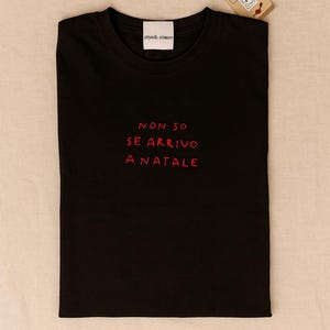Può includere: T-shirt nero con testo ricamato rosso "NON SO SE ARRIVO A NATALE". La maglietta ha un girocollo e una piccola etichetta in alto con "cryedi.cimon". Un piccolo cartellino con una faccia di cartone animato è attaccato.