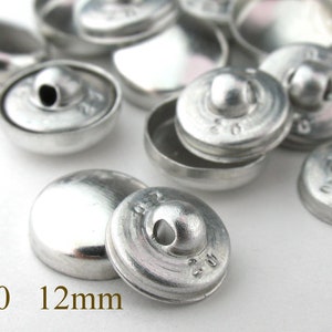 Op de afbeelding: Een close-up van een stapel zilveren metalen knopen, elk met een verhoogd midden en een #20 markering. De knopen zijn 12 mm in diameter.