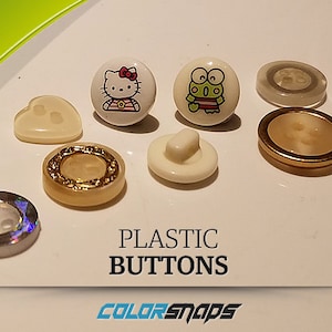May include: Assortment of plastic buttons in various colours and designs, including a heart, a smiley face, a frog, Hello Kitty, and a gold button with a decorative border. The buttons are on a white surface with the text "PLASTIC BUTTONS" and "COLORSNAPS" below.