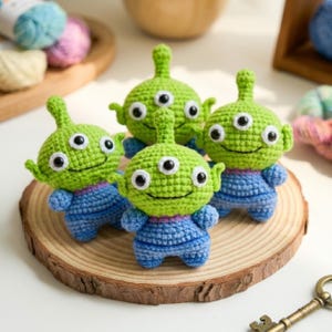 May include: Four small, handmade crochet figures featuring bright green heads with three black and white eyes, pointed ears, and small antennae. Each figure wears a blue crocheted outfit with a simple stitched smile, arranged together on a round wooden base.