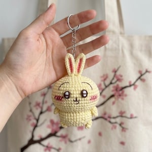 May include: A pale yellow crocheted rabbit keychain featuring long ears with pink centers, embroidered dark brown eyes with white highlights, a small mouth, and pink stitched blush marks on the cheeks, attached to a silver metal split ring.