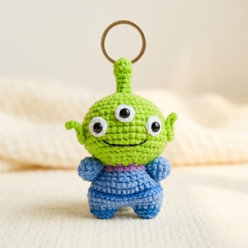 May include: A crochet keychain featuring a small, round, lime green alien character with three black eyes, pointed ears, and a small antenna on top. The figure wears a blue textured outfit and is attached to a circular gold-toned metal ring.