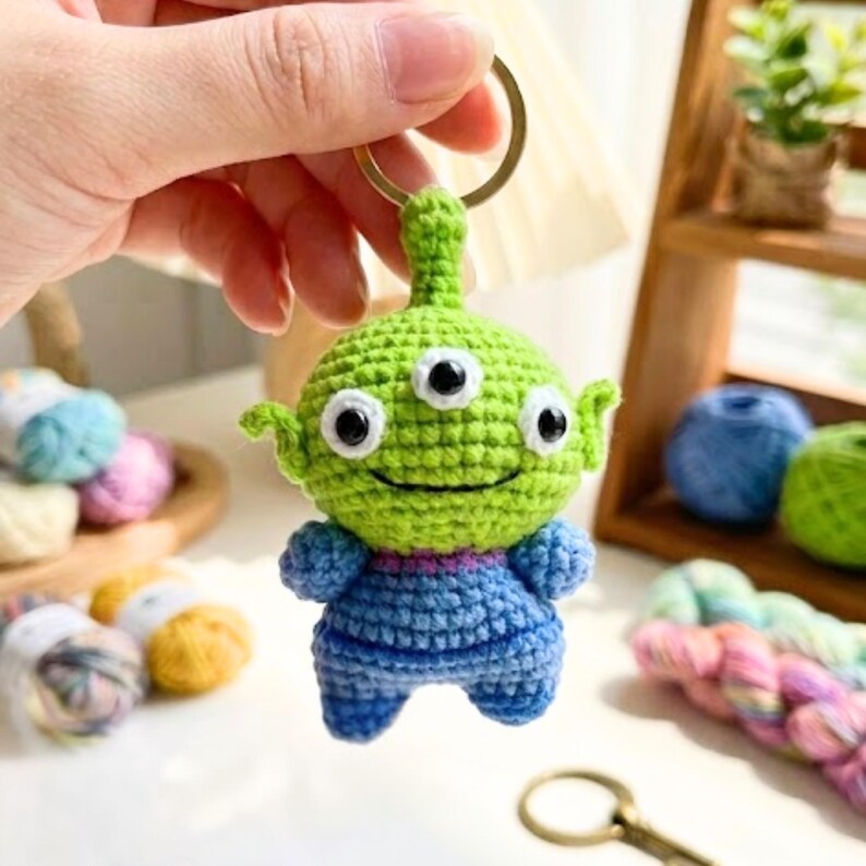 May include: A crochet keychain featuring a green three-eyed alien character with a round head, pointed ears, and a blue body. The figure is crafted with visible yarn stitches and is attached to a circular metal ring for hanging purposes.