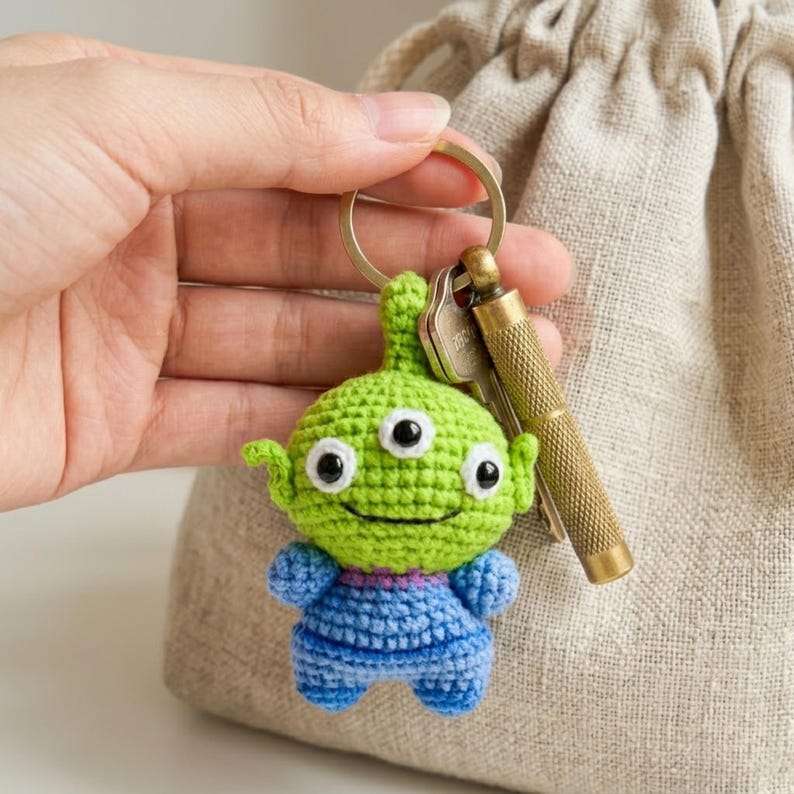 May include: A crochet keychain featuring a small green three-eyed alien character with a blue shirt, attached to a gold-toned metal ring along with a silver key and a textured brass-colored cylindrical tool.