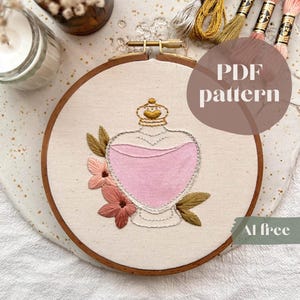 Puede incluir: Arte de aro bordado con un diseño de botella de poción rosa con una corona dorada, rodeada de flores rosas y marrones y hojas verdes. El texto "PDF pattern" es visible. El diseño está sobre una tela color crema.