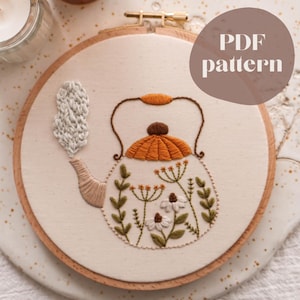 Può includere: Ricamo di un disegno di teiera in un cerchio di legno. La teiera è arancione e marrone, con vapore bianco e dettagli floreali. Il testo "PDF pattern" è in un cerchio marrone.